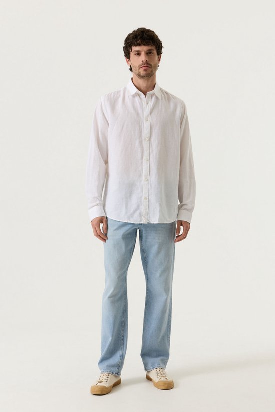 Man met krullend haar, van voren gezien, draagt een wit linnen overhemd met lange mouwen en lichtblauwe jeans.