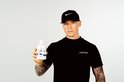 Jonge man met tatoeages, draagt een zwarte pet en een zwart T-shirt, houdt een flesje PROBIOTICS omhoog.