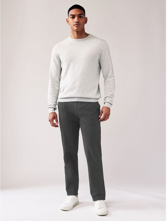 Model draagt een grijze chinobroek met rechte pasvorm, grijze trui en witte sneakers.