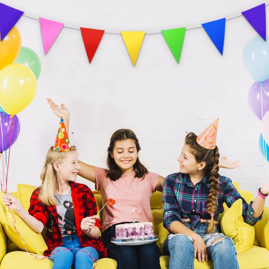 Trois filles portant des chapeaux d'anniversaire et des ballons sur un canapé jaune, avec des fanions colorés.