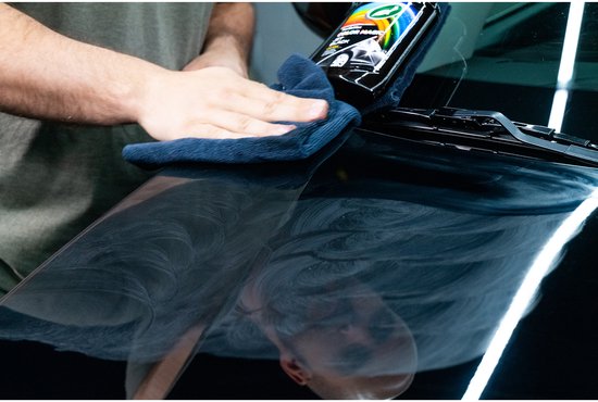 Een man poetst de motorkap van een zwarte auto met een donkere microvezeldoek, de fles wax staat erop.