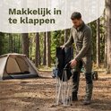 Man in bos vouwt zwarte klapstoel op. Tent en campinguitrusting op de achtergrond.