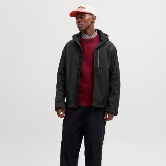 Man met zwarte softshell jas met capuchon, open rits, rood gebreide trui en geruit overhemd, staand.