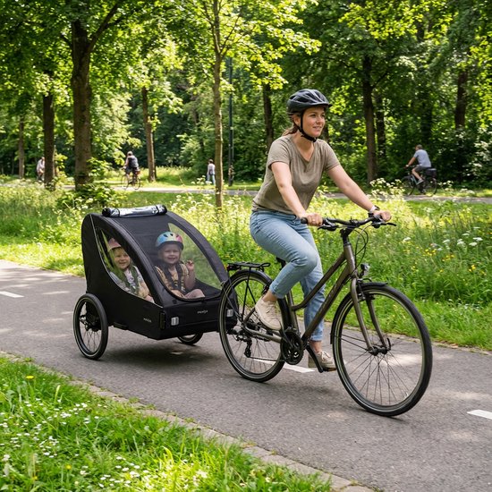 Een vrouw met helm rijdt op een fiets met een zwarte fietskar met twee kinderen erin.