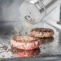 Zilverkleurige kruidenstrooier strooit kruiden op een gegrilde hamburger.