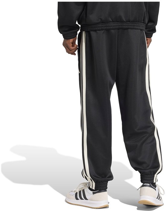 Achterkant van een man in zwarte trainingsbroek met witte strepen en Adidas logo, gedragen met witte sneakers.