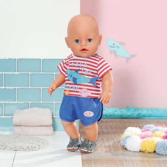 Baby Born pop staand, met rood-wit gestreept shirt met haai en blauwe broek. Draagt grijze haaienschoentjes.