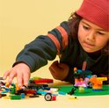 Jongen met rode muts, bouwt aan LEGO creaties op groene bouwplaat, met LEGO stenen.
