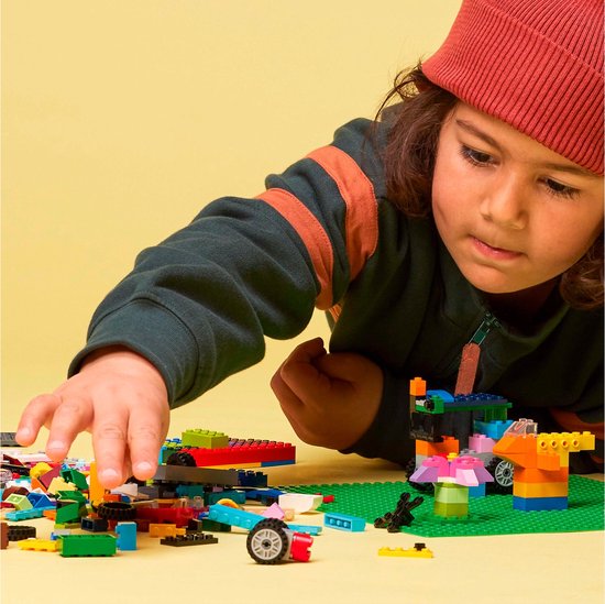 Jongen met rode muts, bouwt aan LEGO creaties op groene bouwplaat, met LEGO stenen.