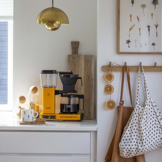 Felgeel Moccamaster koffiezetapparaat met glazen kan gevuld met koffie, naast keukengerei en een tas.