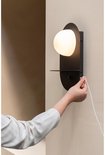 Hand steekt de stekker in een zwarte wandlamp met ovale melkglazen kap en een schapje.