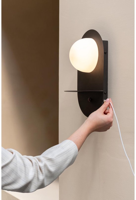Hand steekt de stekker in een zwarte wandlamp met ovale melkglazen kap en een schapje.