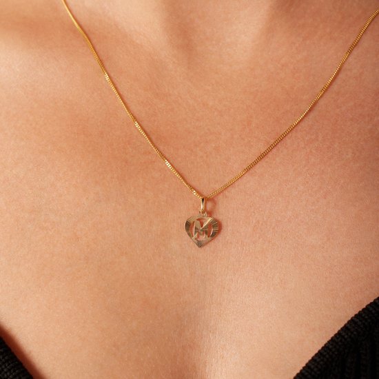Close-up van een gouden hartvormige hanger met de letter M aan een ketting, gedragen op de huid.