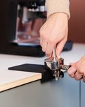 Handen die een portafilter tappen op een zwarte tampermat naast een espressomachine.