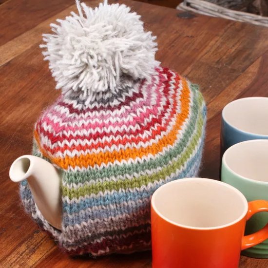Theemuts met regenboogstrepen en een grijze pompon, naast gekleurde mokken.