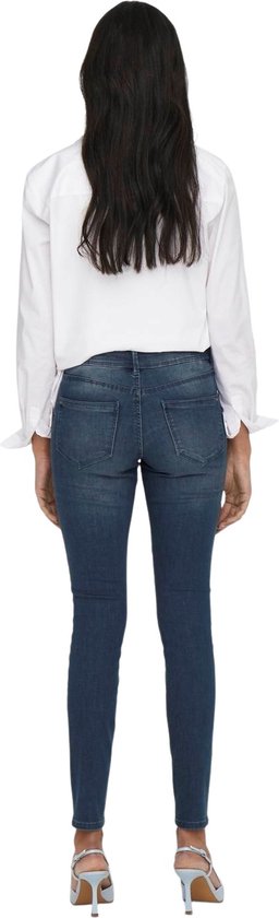 Model in een donkerblauwe skinny jeans met lichte slijtageplekken en een witte blouse.