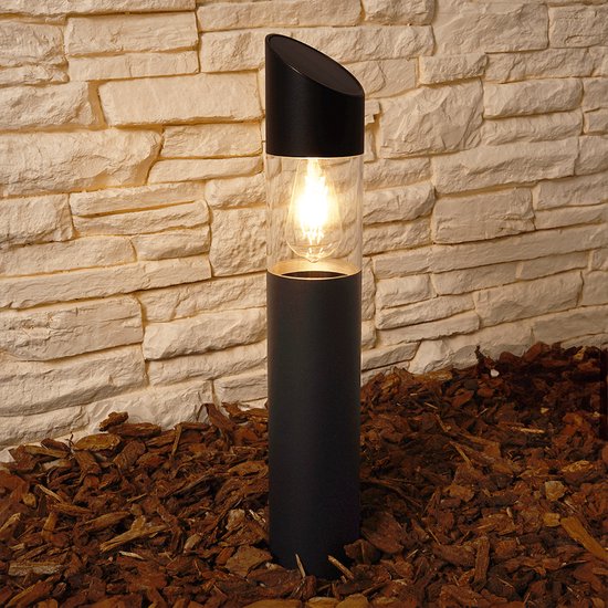 Zwarte staande tuinlamp met verlichting, doorzichtige cilinder en schuin aflopende zwarte kap. Staat op boomschors.