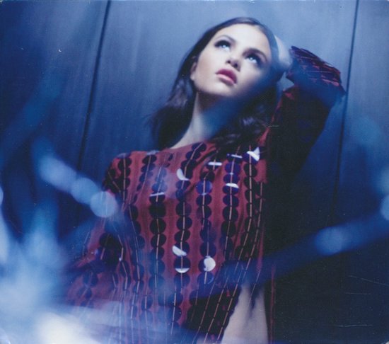 Selena Gomez in een rode blouse met cirkelpatroon en ronde zilveren pailletten, kijkt omhoog met haar hand achter haar hoofd.