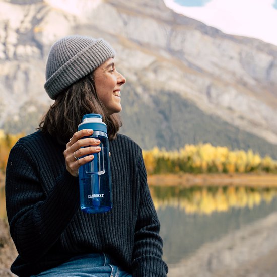 Une femme portant un bonnet gris et un pull bleu foncé, assise dans la nature, tient une gourde Kambukka bleue avec paille.