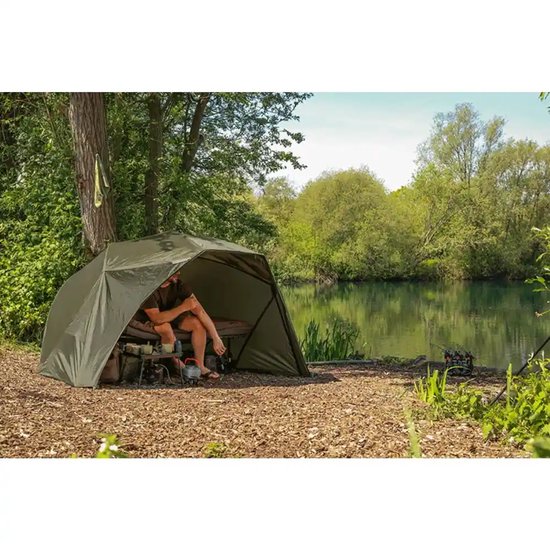 Een olijfgroene visbrolly op de oever, met een man die erin zit, een bedchair en spullen.