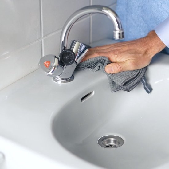 Une main avec un chiffon en microfibre gris polit un lavabo blanc autour d'un robinet.