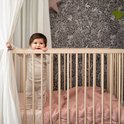 Baby staat in een ledikant. Draagt een beige longsleeve broekpak.
