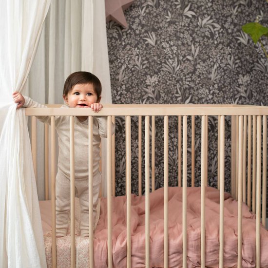 Baby staat in een ledikant. Draagt een beige longsleeve broekpak.