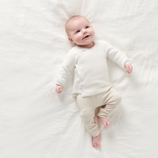Baby in een wit gebreid vestje met omslagsluiting, liggend op een witte sprei.