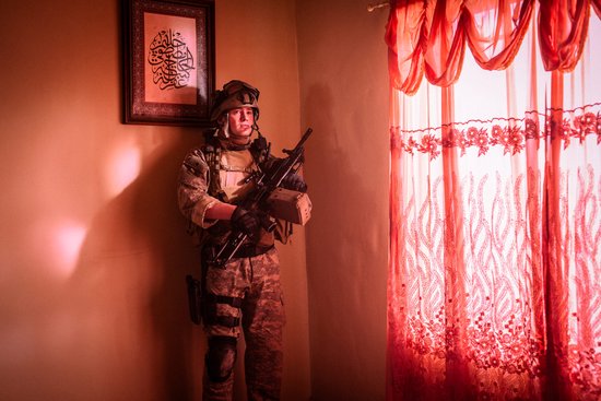 Soldaat in camouflageuniform met helm en wapen staat in een kamer met decoratieve rode gordijnen.