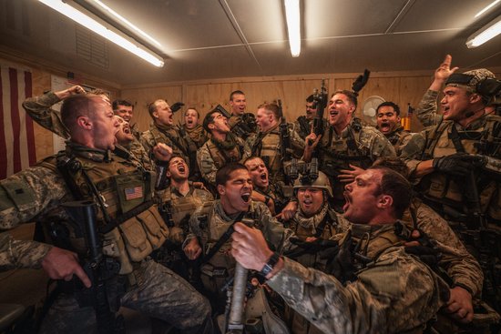 Een groep lachende en schreeuwende militairen in gevechtskleding, sommigen met wapens, in een kamer.