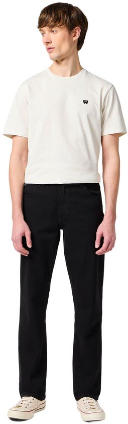 Man in wit T-shirt met logo en zwarte jeans, staand.