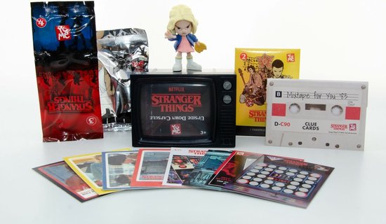 Figurine d'Eleven de Stranger Things, entourée de pochettes d'emballage, de cartes, d'une cassette et d'un écran de télévision rétro.