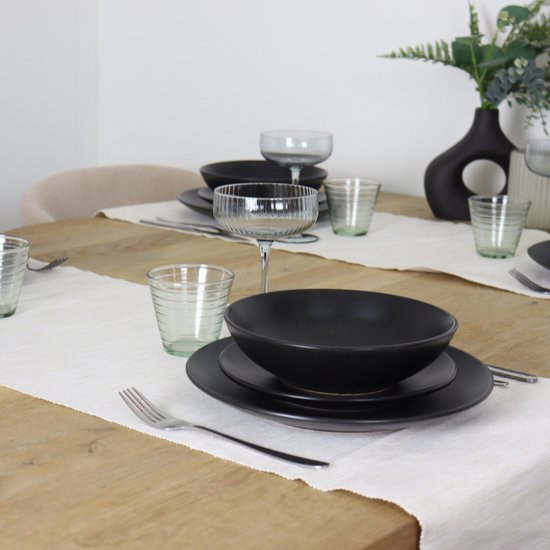 Een tafel gedekt met matzwarte borden, glazen en bestek, met een vaas met groen gebladerte op de achtergrond.