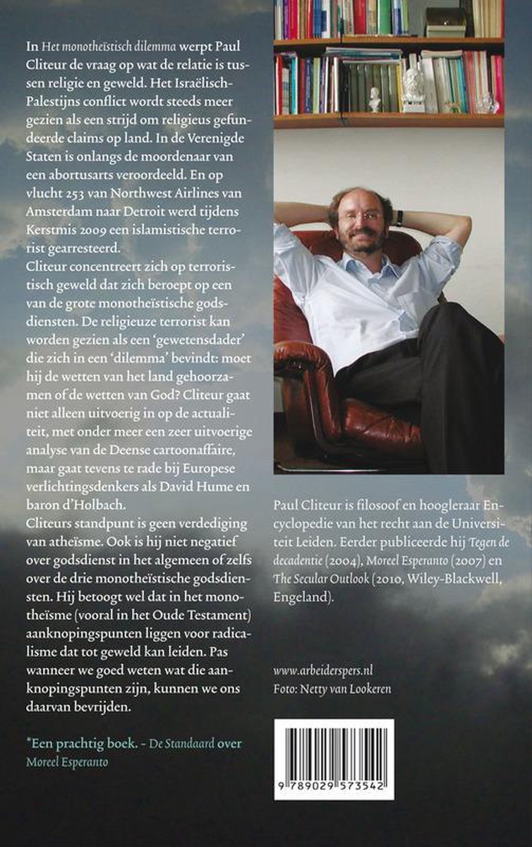 Een man met bril en baard zit ontspannen in een stoel, met zijn armen achter zijn hoofd. Boekenplanken vol boeken op de achtergrond.