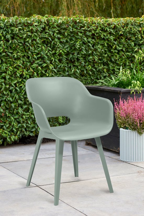 Une seule chaise de jardin vert vintage sur une terrasse carrelée, avec une haie verte et des plantes fleuries en arrière-plan.