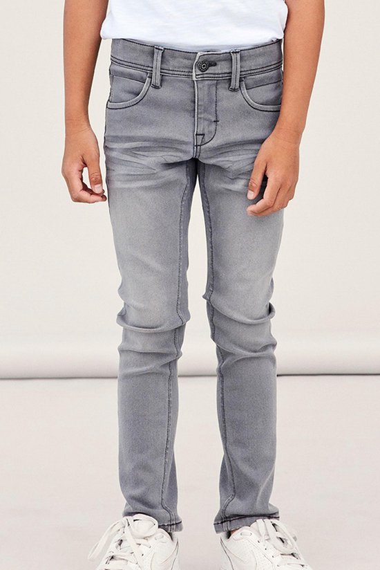 Jongen draagt een grijze, aansluitende jeans met gedetailleerde stiksels en witte sneakers.