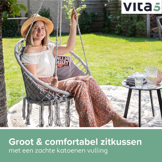 Grijze macramé hangstoel met franjes, waarin een vrouw met strooien hoed en bloemenprint broek zit.