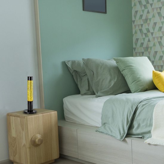 Een gouden glitter lavalamp staat op een houten nachtkastje naast een bed met groen beddengoed.