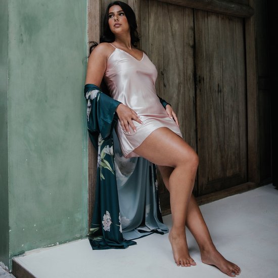 Model met lange zwarte haren leunt tegen een houten deur, draagt een roze satijnen slipdress met een donkergroene kimono met bloemenprint.