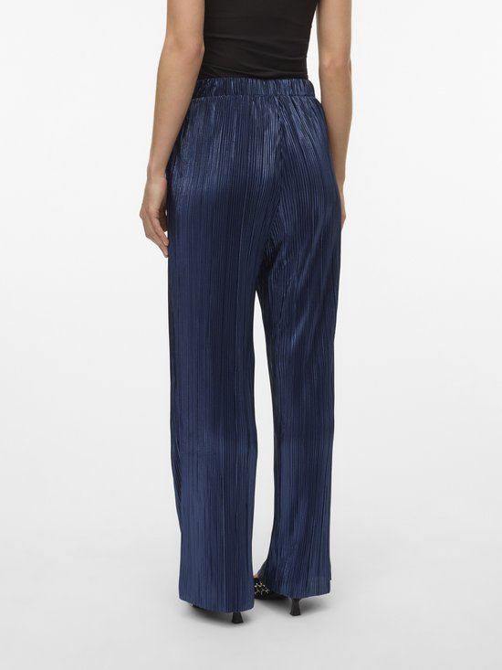 Model draagt een donkerblauwe, glanzende, geplooide broek met wijde pijpen.