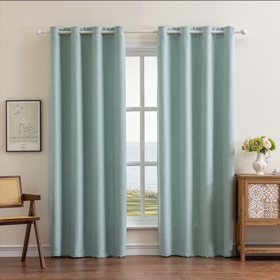 Twee lichtblauwe gordijnen met linnenlook textuur hangen voor een raam met uitzicht op zee. Een houten stoel staat links, een kastje rechts.
