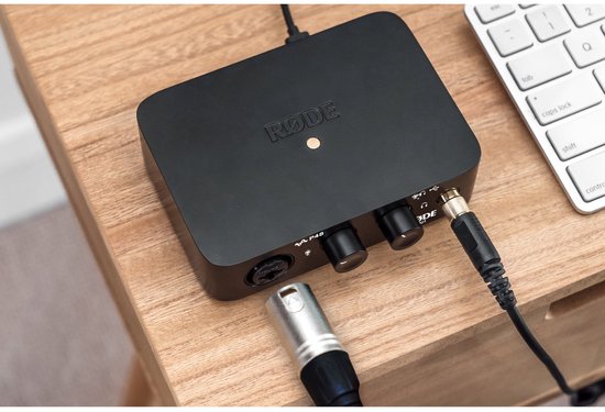 Zwarte RØDE AI-1 audio interface aangesloten op een microfoon en aangesloten op een computer, op een houten bureau.