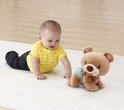 Baby in geel-wit gestreept shirt kruipt op een vloerkleed naar een bruine beer met luier.