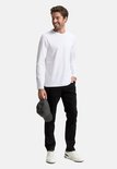 Man in zwarte jeans en witte longsleeve, met pet in hand.