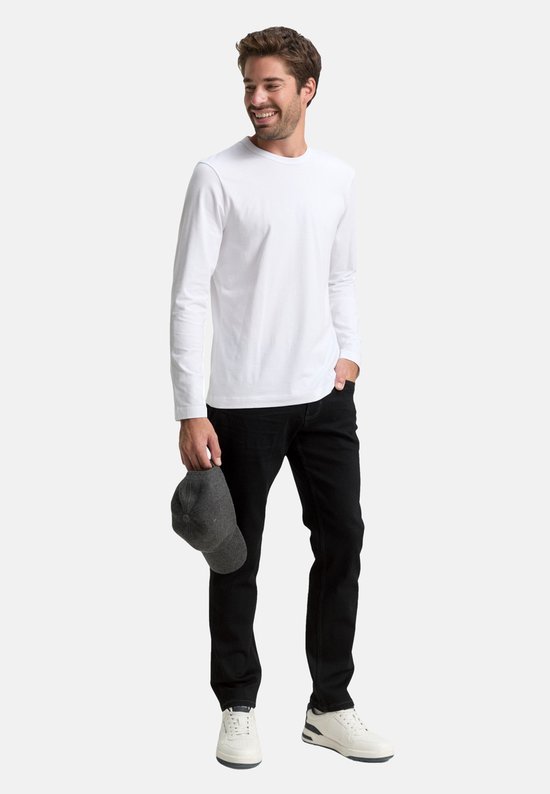 Man in zwarte jeans en witte longsleeve, met pet in hand.