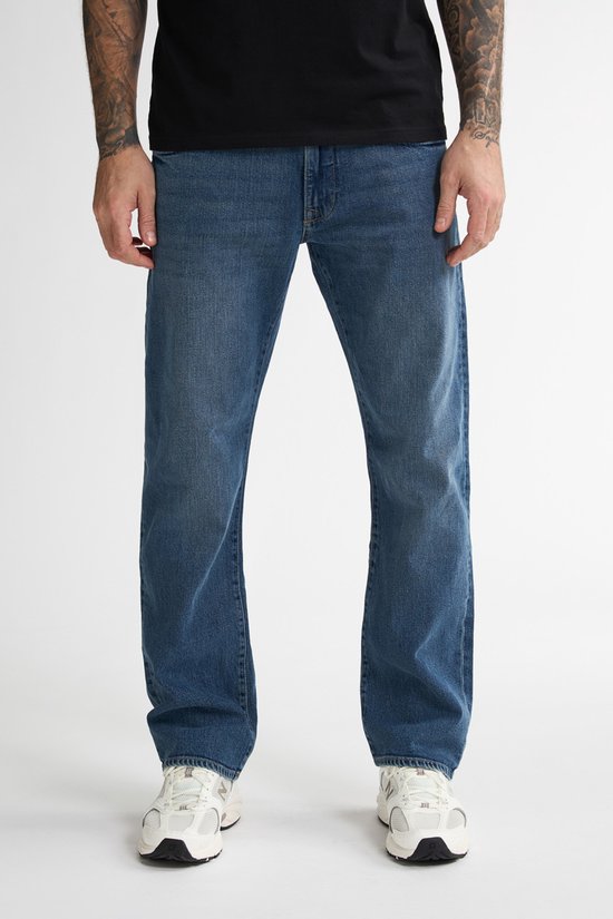 Man draagt losse blauwe jeans en witte sneakers. Voorkant zichtbaar.