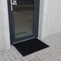 Zwarte ringmat mat bij de ingang van een gebouw, op een grijze steenstrook.