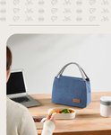 Blauwe lunchtas met touwen handvatten op een bureau, naast een laptop en lunchboxen. Een vrouw met een bril eet een salade.