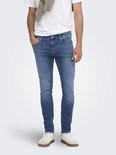 Model in blauwe skinny jeans, wit T-shirt, beige trui en witte sneakers.