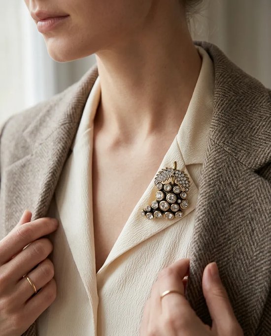 Model draagt een koperen druiventrosbroche met fonkelende zirkonia's op de kraag van een beige blouse.